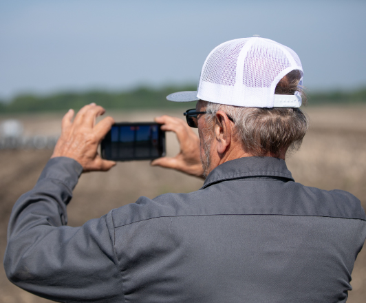 Farmer holding phone up to take a picture of a tractor pulling a planter